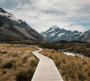 Wooden walkway leads through rugged landscape towards majestic snow-capped mountains.
